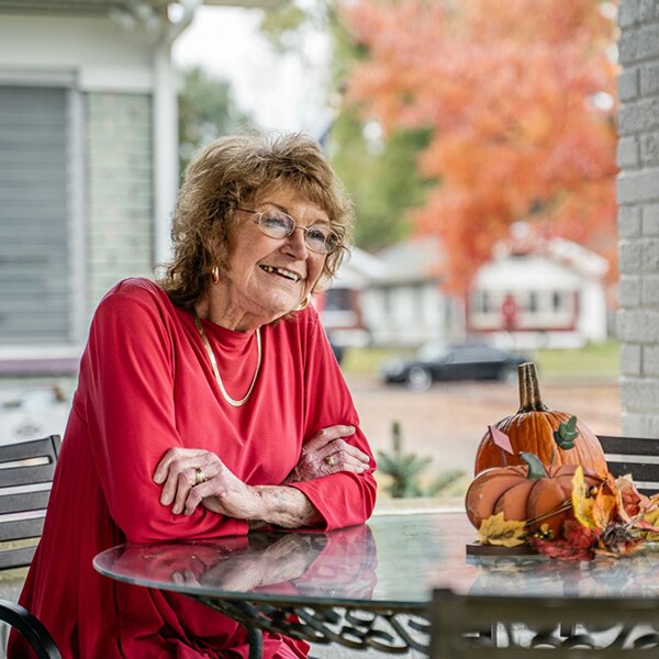 A smiling, seated woman in a red shirt at a patio table outside her home