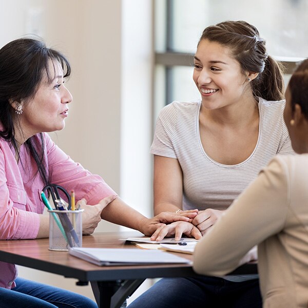 Three women at a table with papers and office supplies in a brightly lit room talking to each other