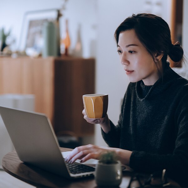 Person working on a laptop at a dining table while holding a coffee mug in a home setting.
