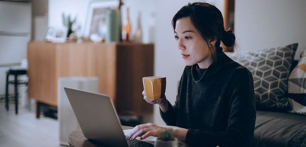 Person working on a laptop at a dining table while holding a coffee mug in a home setting.