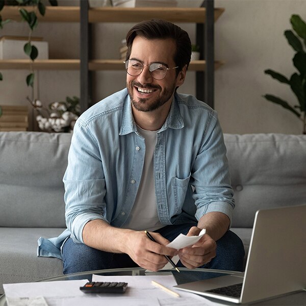 Person seated on a couch reviewing paperwork with a laptop and calculator on a coffee table.