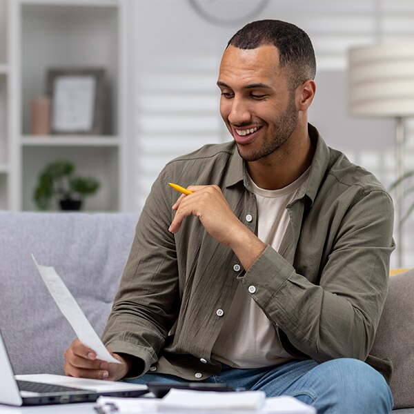 Person seated on a couch reviewing paperwork while holding a pencil, with documents spread out nearby.