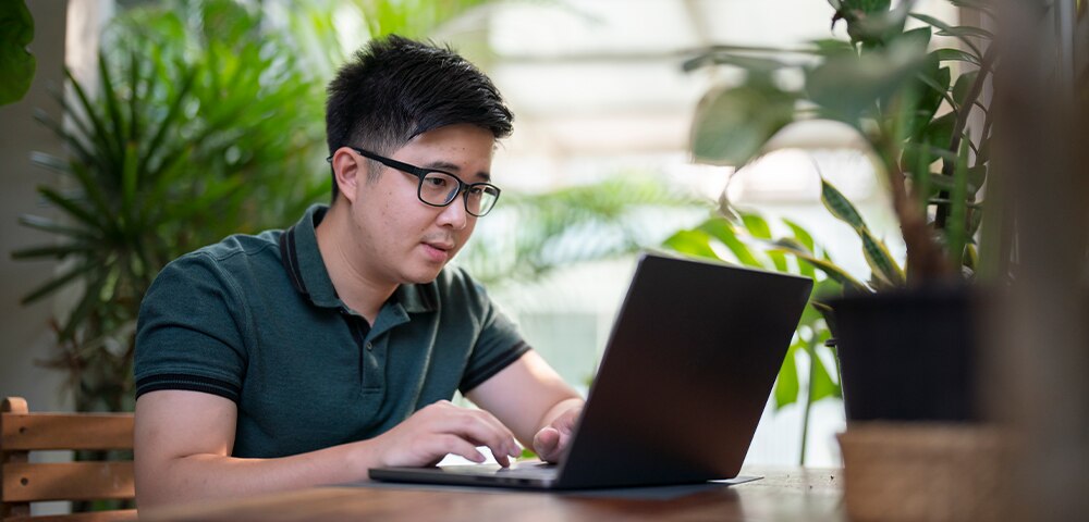Person working on a laptop at a table surrounded by plants, researching information online in a bright indoor workspace.