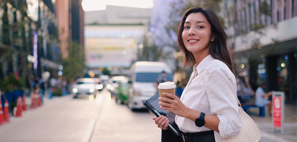 Person holding a takeaway coffee and tablet while standing on a city street.