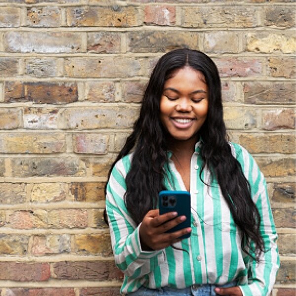 Person standing against a brick wall, wearing a green‑and‑white striped shirt and jeans, holding a smartphone and reading the screen.
