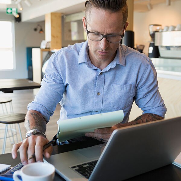 man sitting at table with computer looking at a notepad in hand