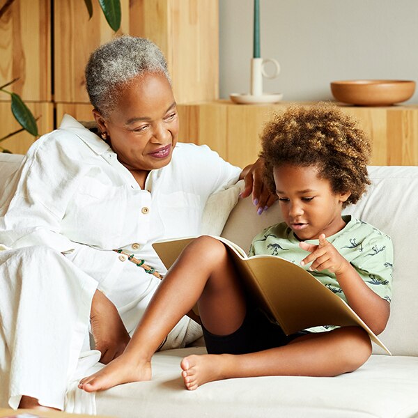 Senior woman looking at grandson reading picture book on sofa.
